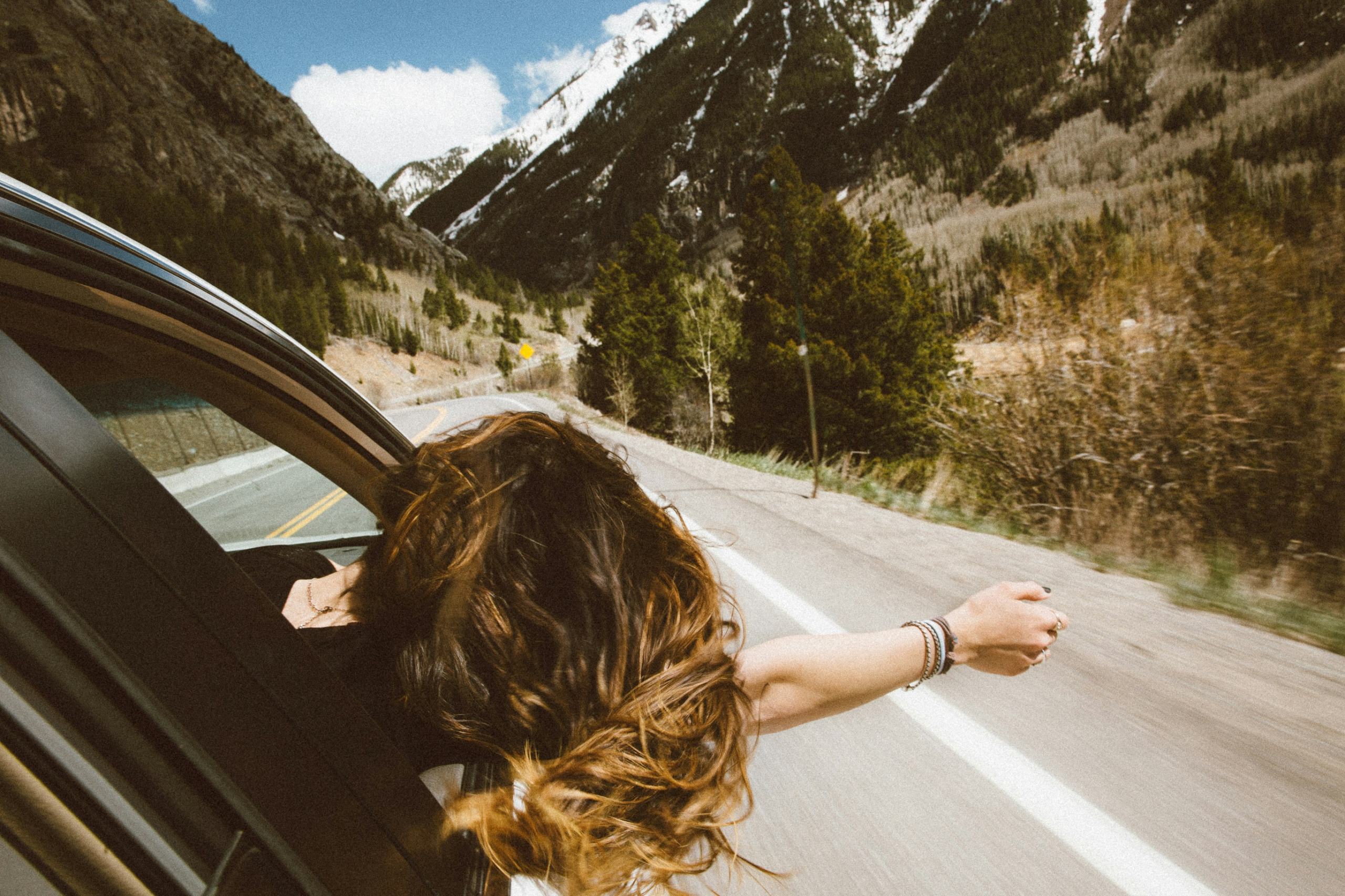 gratitude mistakes - woman leaning out of a moving car along a mountainous road, seen from behind