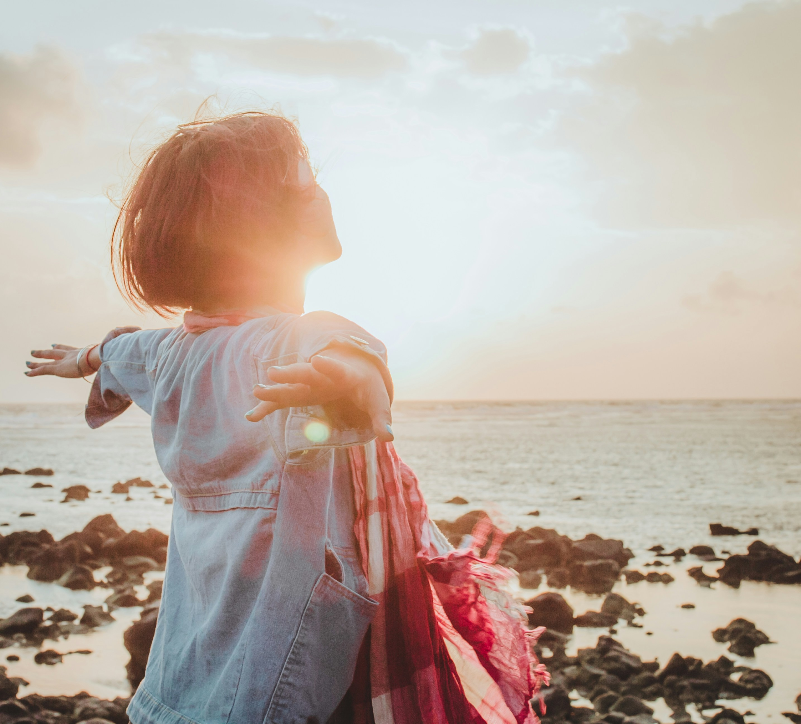gratitude in the midst of chaos - woman facing the sun with arms in the air by the sea