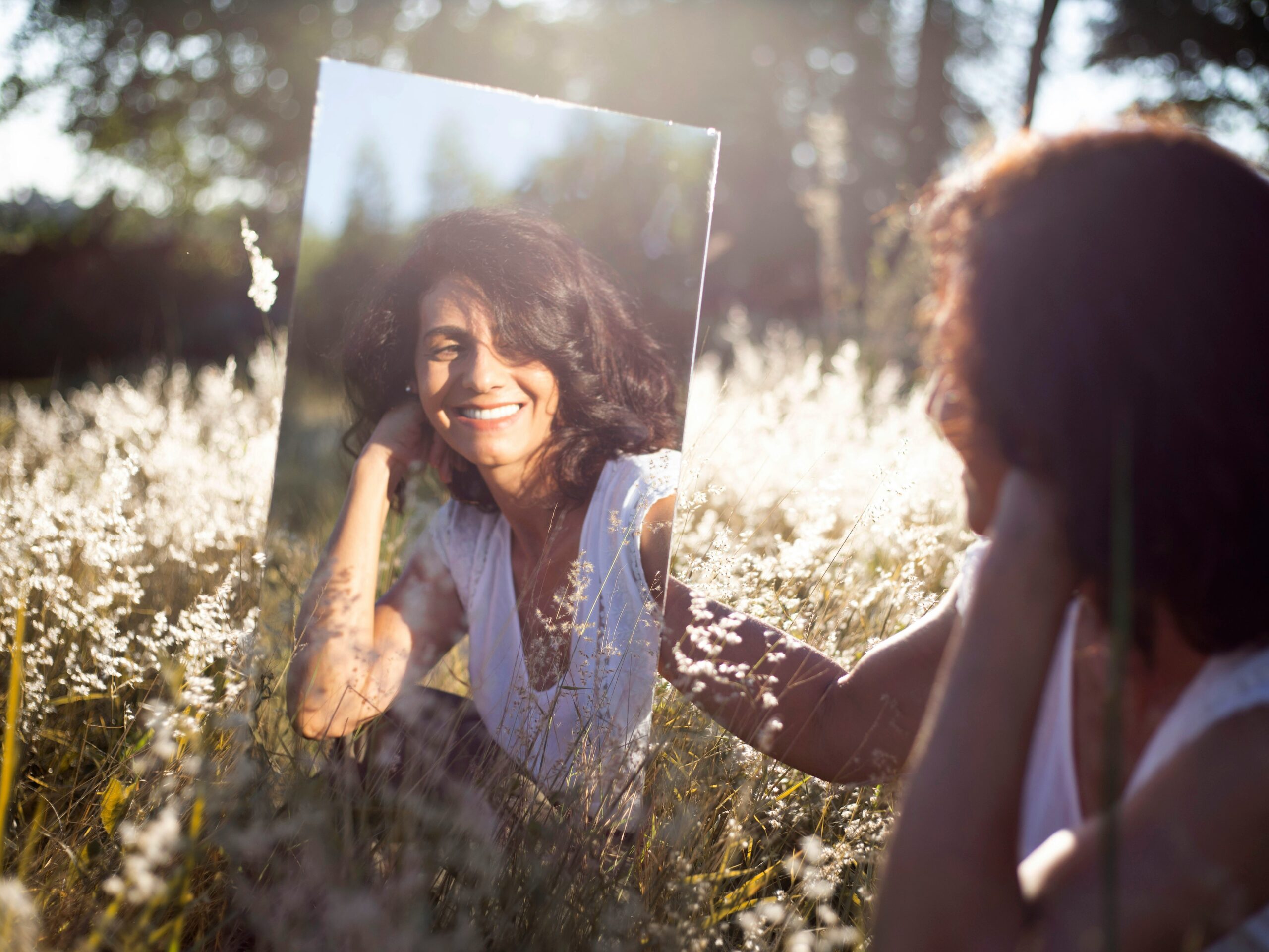 how to stop being so hard on yourself - Woman in a field looking into a mirror and smiling